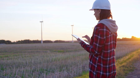 Woman engineer wearing a white protective helmet is taking notes with a tablet computer on a field with wind turbines, as the sun sets. Clean energy and engineering conceptの写真素材