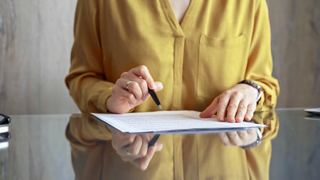 Businesswoman in yellow blouse is signing corporate contract at her office desk in formal white...