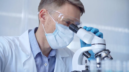 Man scientist with protective glasses, mask and gloves is using microscope in laboratory, portrait...