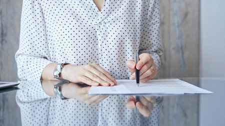 Businesswoman is signing corporate contract at her office desk in formal white blouse with polka...