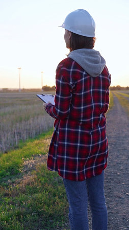 Woman engineer wearing a white protective helmet is taking notes with a clipboard in a field with...