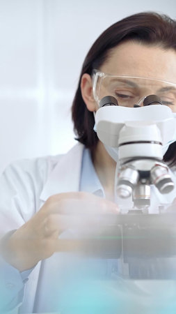 Scientist using microscope in laboratory. Focus on a woman in lab goggles analyzing samples with a...