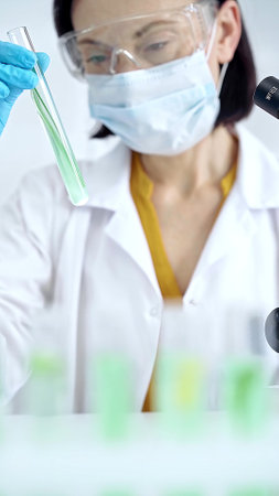 Woman scientist with protective gloves and glasses examining lab tube with a green fluid. Microbiology and science conceptの写真素材