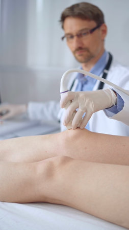 Doctor man with blue protective gloves using ultrasound equipment on the knee of a female patient, analyzing the results on a monitor in the background. Medicineの写真素材
