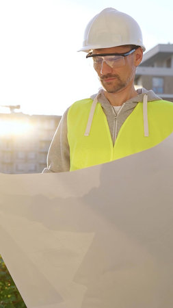 Man an architect or engineer wearing safety hard hat and vest holding blueprint and looking to...
