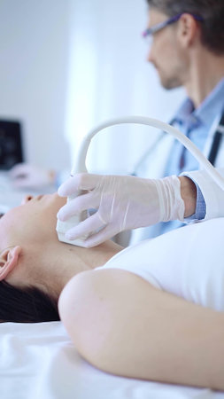 Doctor man wearing white protective gloves is using ultrasound machine to examine neck of female patient in hospital. Medicine, thyroid gland healthの写真素材
