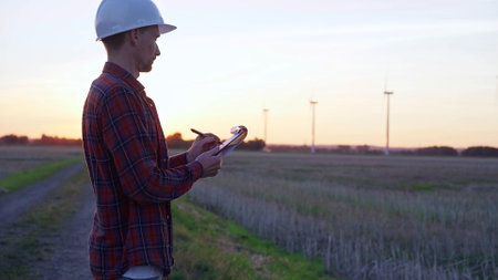 Man engineer wearing a white protective helmet is taking notes with a clipboard in a field with wind turbines, as the sun sets. Clean energy and engineering conceptの写真素材