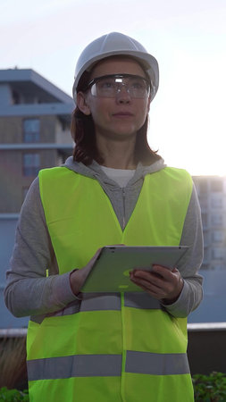 A woman construction engineer with a white hard hat and safety vest is using a digital tablet while...