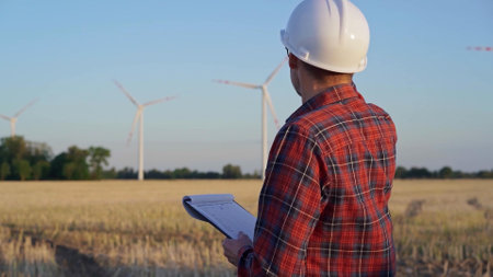 Man engineer, wearing a white protective helmet is taking notes with a clipboard in a field with wind turbines, as the sun sets. Clean energy and engineering conceptの写真素材