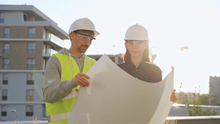 Two architects wearing safety hard hats are holding blueprint and discussing something on construction site at sunrise. Architect and teamwork conceptの写真素材