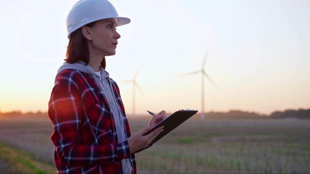 Female engineer taking notes on a clipboard on a field with wind turbines, as the sun sets. Concept of clean energy and engineering auditの写真素材