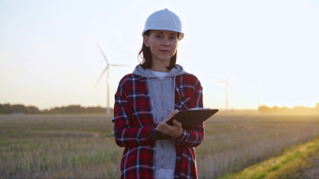 Female engineer is standing straight and keeping clipboard on a field with wind turbines, as the sun sets. Concept of clean energy and engineering auditの写真素材