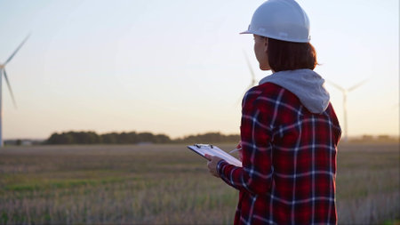 Adult woman engineer wearing white hard hat is taking notes on a clipboard on a field with wind turbines, as the sun sets, back viewの写真素材
