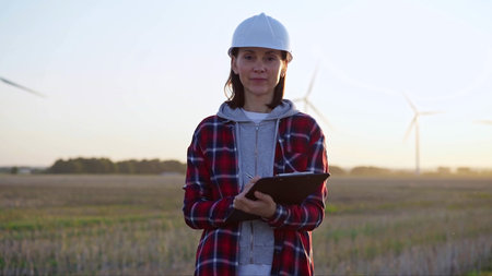 Female engineer is standing straight and keeping clipboard on a field with wind turbines, as the sun...
