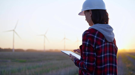 Female engineer taking notes with a tablet computer on a field with wind turbines, as the sun sets. Concept of clean energy and engineering auditの写真素材