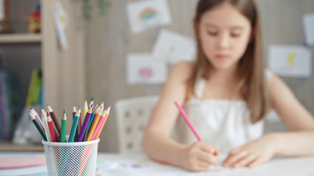 A close-up of colored pencils on the desk, with a creative young girl drawing in the background