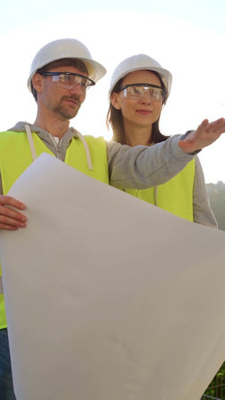Man and woman architects or engineers wearing safety hard hats and vests holding blueprint and discussing something on construction site at sunrise, front vertical view. Architecture conceptの写真素材