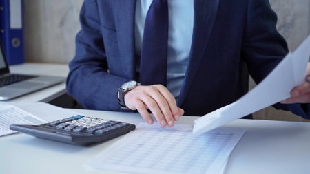 Businessman accountant with blue suit is calculating numbers for a financial audit and taking notes with a silver pen. Taxes and audit conceptの写真素材