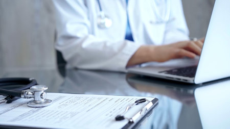Stethoscope in the focus. Doctor woman is using laptop computer in clinic office while sitting at the glass desk in medical modern office. Medicine and health careの写真素材