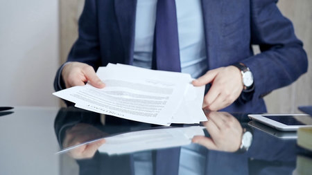 Businessman reviewing documents at office desk. Close-up of a professional mans hands examining paperwork with pen and tablet in sight. Business people conceptの写真素材