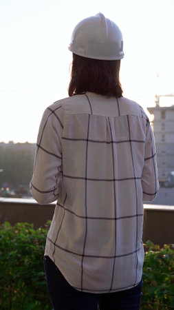 Woman a construction engineer wearing white checked blouse and hard hat is using tablet computer while inspecting a building site at sunset, back and vertical view. Architecture conceptの写真素材