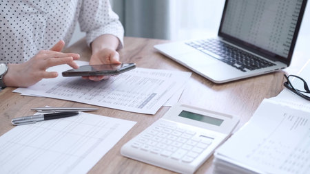 Close-up of an adults hands with smartphone, calculator, and documents on a desk in office....