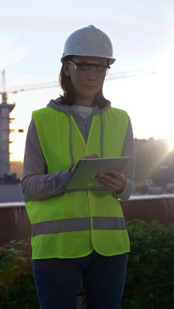 Woman engineer wearing safety hard hat and vest holding digital tablet while inspecting a construction site at sunrise in early morning, front vertical view. Architecture conceptの写真素材