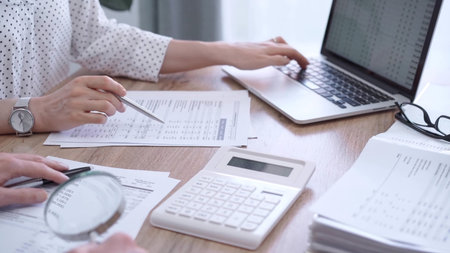 Female bookkeeper or auditor is working on finances papers with a colleague, using calculator and...