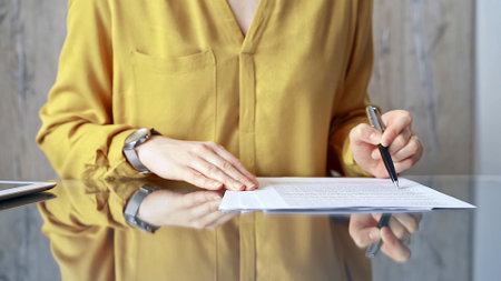 Businesswoman in yellow analyzing document at desk. Close-up of a professional auditor or lawyer reviewing a lengthy paper report in office setting. Business people conceptの写真素材