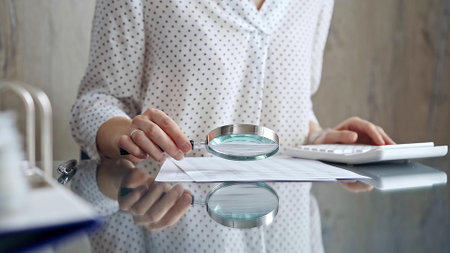 Auditor woman analyzing financial documents with magnifying glass and calculator at her office desk in formal white blouse with polka dots. Business people conceptの写真素材