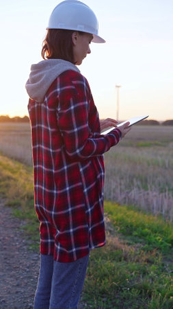 Woman engineer wearing a white protective helmet is taking notes with a clipboard in a field with wind turbines, as the sun sets. Clean energy and engineering conceptの写真素材