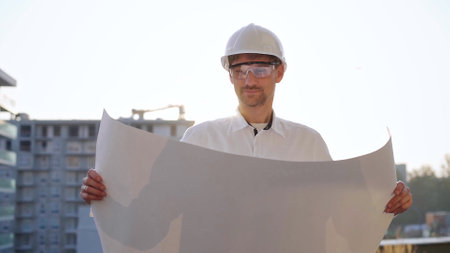 Male construction site manager, wearing safety helmets and glasses, is holding blueprints and inspecting a building site during sunrise or sunset, front side view. Architecture and engineeringの写真素材