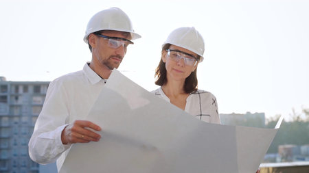 Two architects, wearing safety helmets and glasses, are examining blueprints at a construction site...