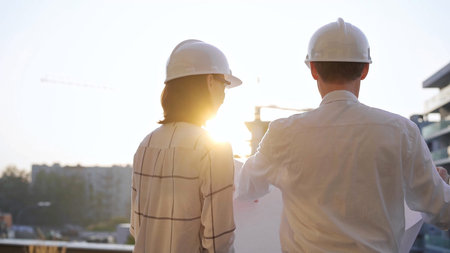 Two architects, wearing hard hat and safety glasses, are examining blueprints at a construction site...