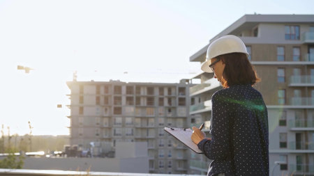 Woman architect wearing a hard hat is taking notes with a tablet computer while inspecting a building construction site at sunsetの写真素材