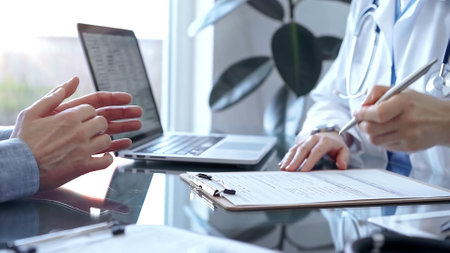 Doctor woman wearing white medical coat is consulting patient in clinic office while both are sitting at the glass desk. Physician is listening and making notes. Medicine conceptの写真素材