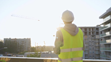 Male engineer with white hard hat and safety vest is taking notes with a tablet computer while...