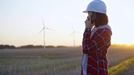 Woman engineer talking on the phone in a field with wind turbines, as the sun sets. Concept of clean...