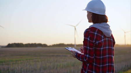 Adult woman engineer wearing white cask is taking notes on a clipboard on a field with wind...