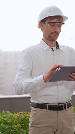 Man a construction engineer or architect, wearing a white shirt and helmet, is using a digital...