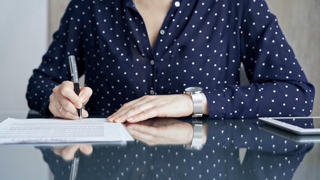 Businesswoman is signing corporate contract at her office desk in formal dark blue blouse with polka...