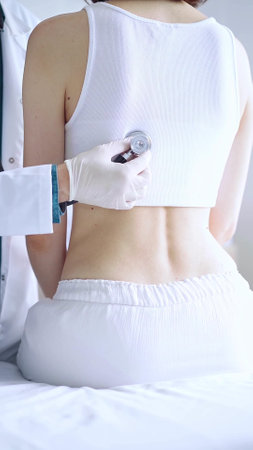 Doctor and patient. Physician wearing white medical gloves is using stethoscope examining woman lungs from the back side in clinic. Medicine and health careの写真素材