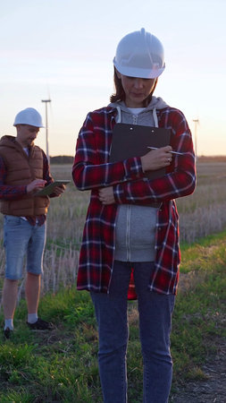 Woman engineer is standing straight and taking notes with a clipboard in a field with wind turbines,...