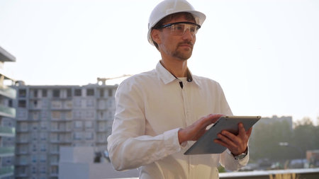 Male construction engineer or architect wearing white shirt and hard hat is using digital tablet...