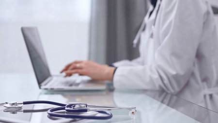 Tablet computer and stethoscope are lying on the glass table while doctor woman is using laptop...