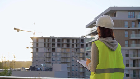 Female construction engineer with a hard hat and safety vest is writing on a clipboard while inspecting a construction site in the evening at sunsetの写真素材