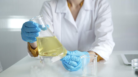 Scientist, wearing blue protective gloves, is holding a beaker with yellow liquid while sitting at...