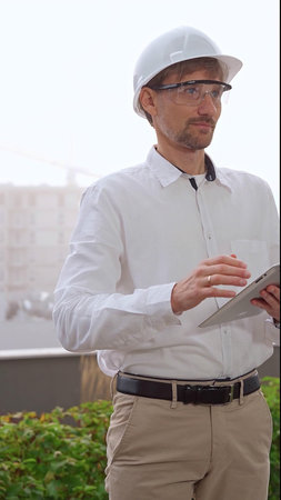 Male architect, wearing a white shirt and helmet, is using a digital tablet while inspecting a...