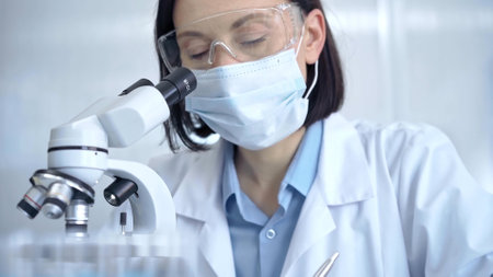 Female scientist in lab goggles, protective mask and gloves is using microscope in laboratory. Science conceptの写真素材