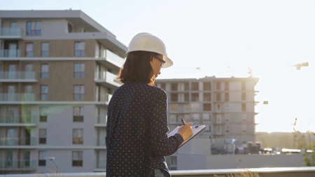 Woman architect wearing a hard hat is taking notes with a tablet while inspecting a building construction site at sunsetの写真素材